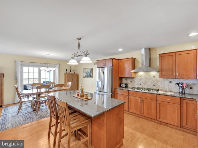 kitchen with appliances with stainless steel finishes, light wood-type flooring, brown cabinets, light stone countertops, and wall chimney exhaust hood