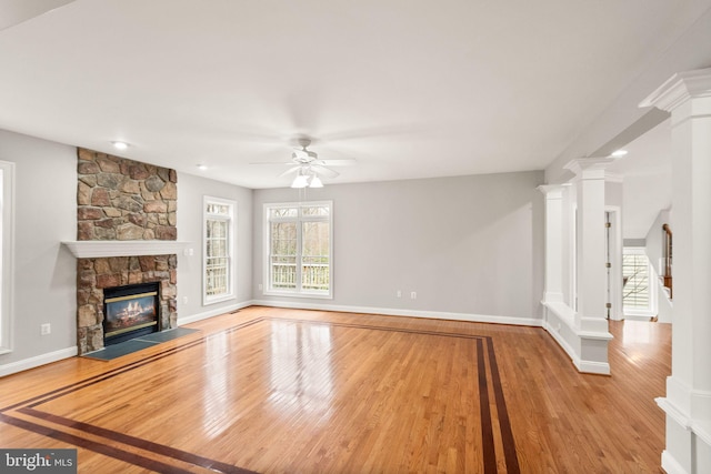 unfurnished living room with ornate columns, ceiling fan, a stone fireplace, wood finished floors, and baseboards