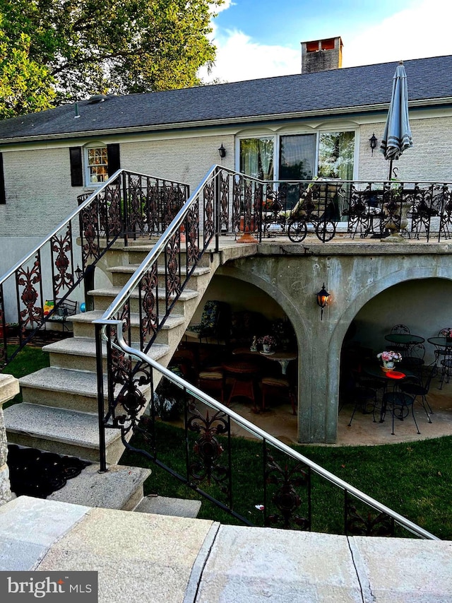 back of property featuring a patio, brick siding, a shingled roof, stairway, and a chimney