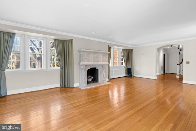 unfurnished living room featuring arched walkways, crown molding, a fireplace with raised hearth, light wood-style floors, and baseboards