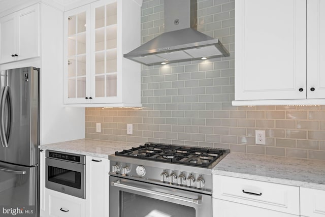 kitchen featuring stainless steel appliances, wall chimney range hood, glass insert cabinets, and tasteful backsplash