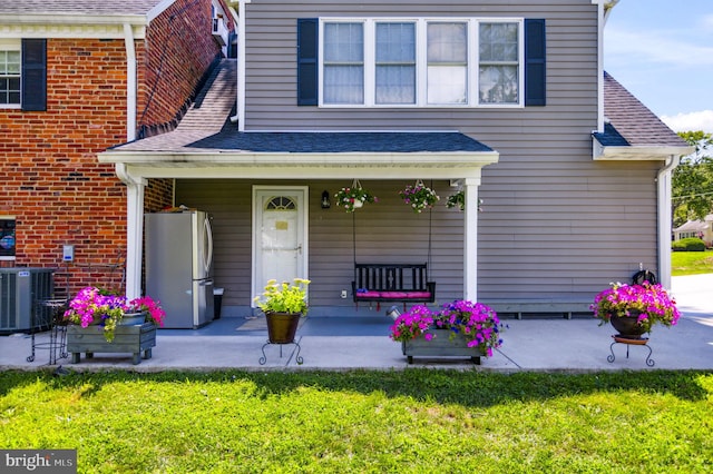 doorway to property with central AC unit, a porch, a lawn, and a shingled roof