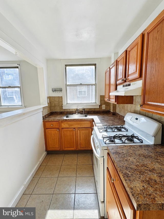 kitchen with white gas stove, under cabinet range hood, a sink, brown cabinets, and dark countertops