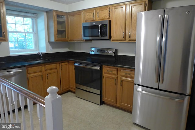 kitchen with brown cabinets, appliances with stainless steel finishes, glass insert cabinets, a sink, and dark stone countertops