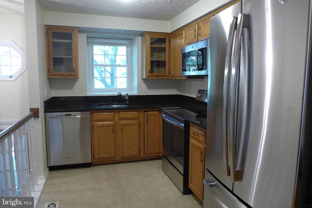 kitchen featuring a textured ceiling, a sink, appliances with stainless steel finishes, brown cabinetry, and dark countertops