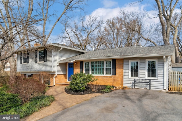 tri-level home featuring brick siding, fence, driveway, roof with shingles, and a chimney