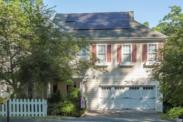 view of front of house featuring driveway, a garage, a shingled roof, fence, and roof mounted solar panels