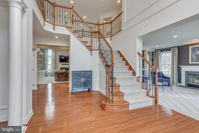 staircase with a high ceiling, wood finished floors, baseboards, ornamental molding, and a glass covered fireplace