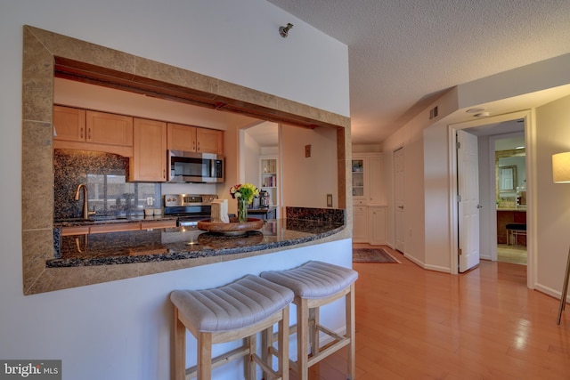 kitchen with light wood-style flooring, a peninsula, appliances with stainless steel finishes, dark stone counters, and a kitchen bar