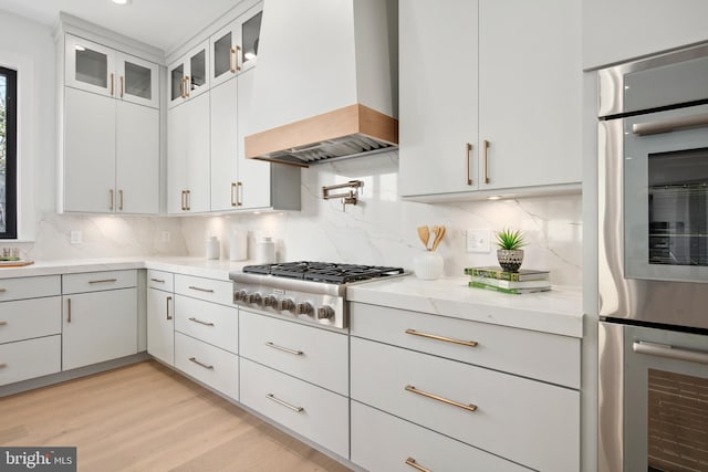 kitchen featuring light wood finished floors, custom range hood, stainless steel appliances, and decorative backsplash