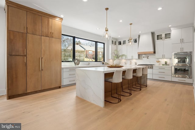 kitchen featuring a breakfast bar area, light wood-style flooring, stainless steel appliances, custom exhaust hood, and glass insert cabinets