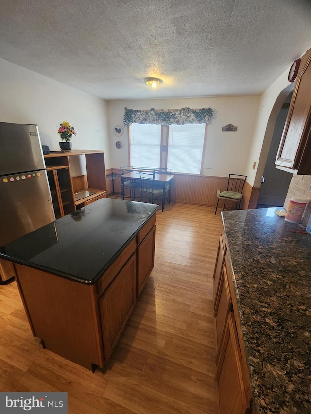 kitchen featuring arched walkways, light wood-style flooring, wainscoting, freestanding refrigerator, and brown cabinets