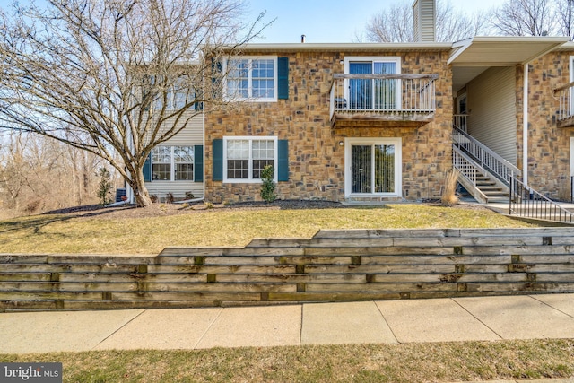 back of house with a balcony, stairs, stone siding, a lawn, and a chimney