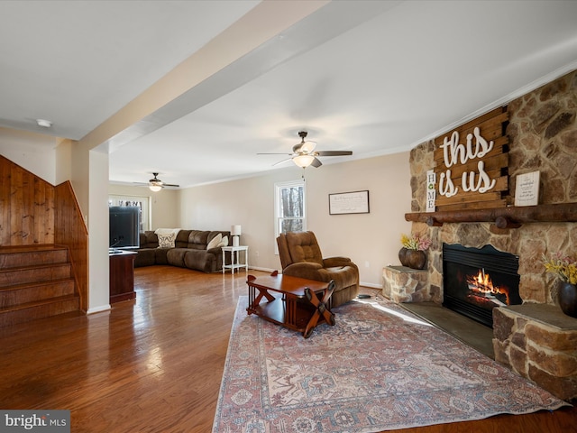 living area with baseboards, stairway, a stone fireplace, wood finished floors, and a ceiling fan