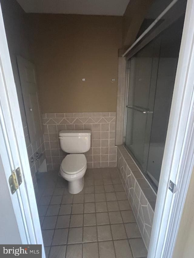 full bathroom with toilet, tile walls, wainscoting, tile patterned flooring, and enclosed tub / shower combo