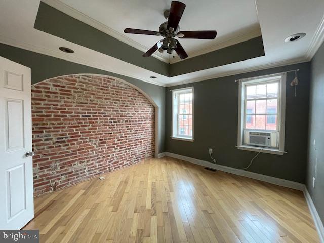 unfurnished room with a tray ceiling, cooling unit, brick wall, and light wood finished floors