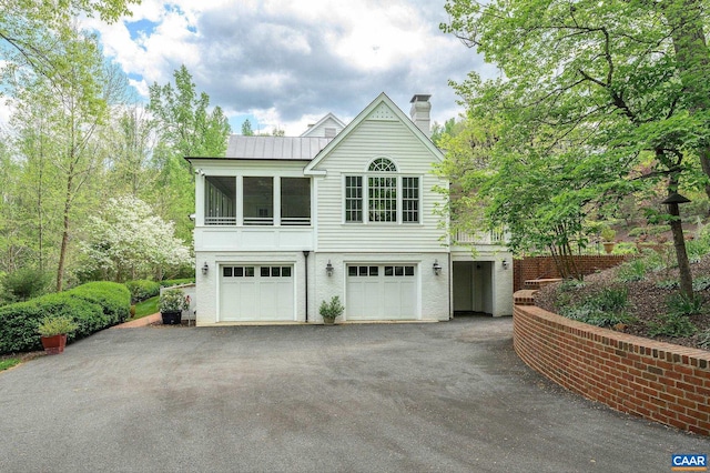 exterior space featuring aphalt driveway, a chimney, a sunroom, metal roof, and a garage