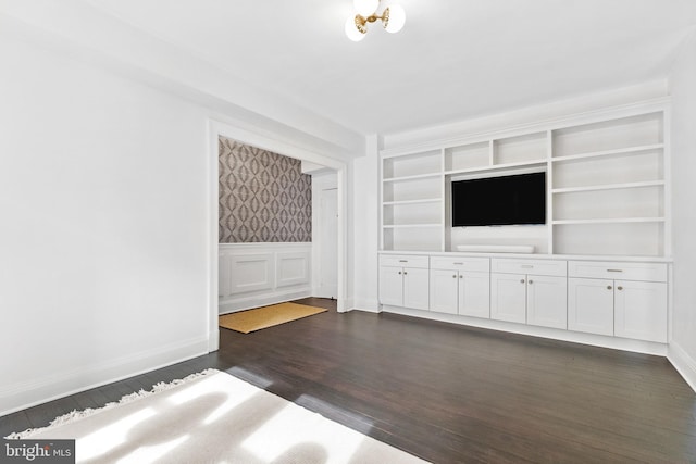 unfurnished living room featuring built in shelves, a wainscoted wall, dark wood-type flooring, baseboards, and wallpapered walls