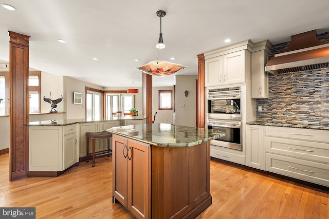 kitchen with light wood-type flooring, wall chimney range hood, double oven, and dark stone counters