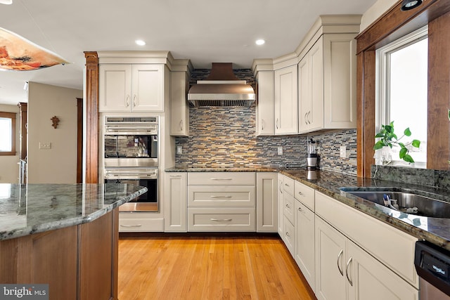 kitchen with stainless steel appliances, backsplash, dark stone counters, light wood-type flooring, and wall chimney exhaust hood