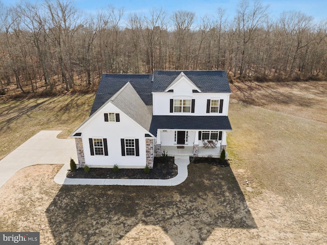 traditional-style house featuring a porch, concrete driveway, stone siding, roof with shingles, and a view of trees