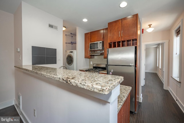 kitchen with a peninsula, appliances with stainless steel finishes, tasteful backsplash, brown cabinetry, and washer / dryer