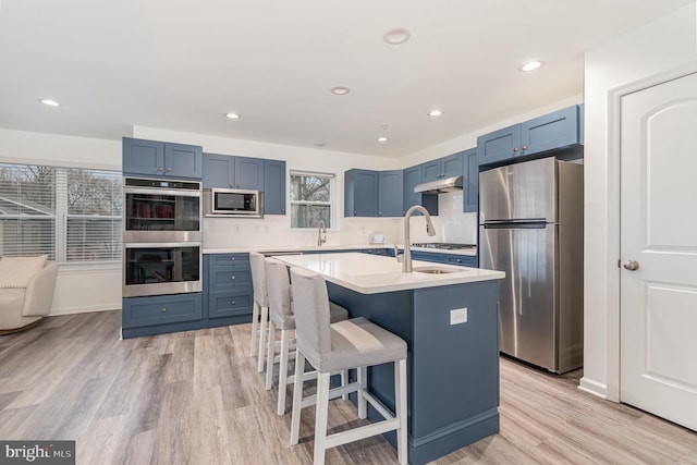 kitchen with a center island with sink, blue cabinetry, stainless steel appliances, light countertops, and light wood-style floors