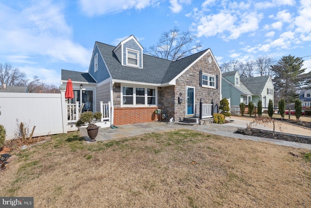 view of front facade with a patio, stone siding, roof with shingles, fence, and a front yard