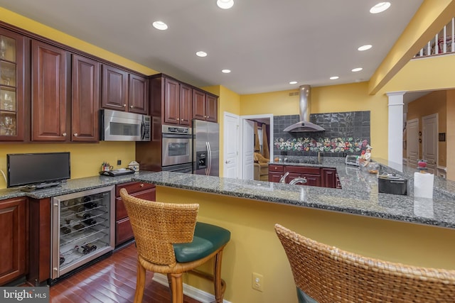 kitchen with beverage cooler, decorative columns, appliances with stainless steel finishes, a kitchen breakfast bar, and wall chimney range hood