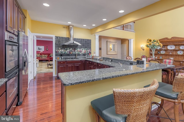 kitchen featuring stainless steel appliances, ornate columns, dark wood-type flooring, ventilation hood, and a kitchen breakfast bar