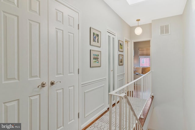 hall featuring wainscoting, visible vents, a decorative wall, and an upstairs landing