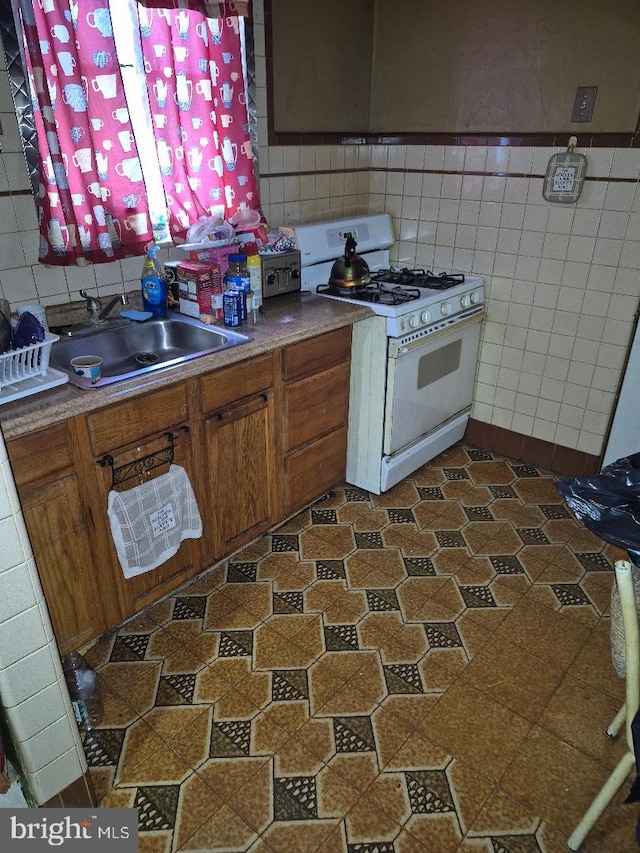 kitchen with white gas stove, brown cabinetry, a sink, and tile walls