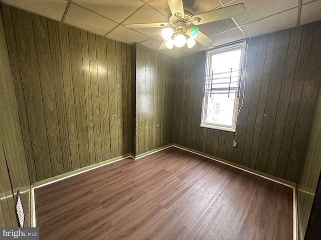 empty room with baseboards, a ceiling fan, a drop ceiling, wood finished floors, and wood walls