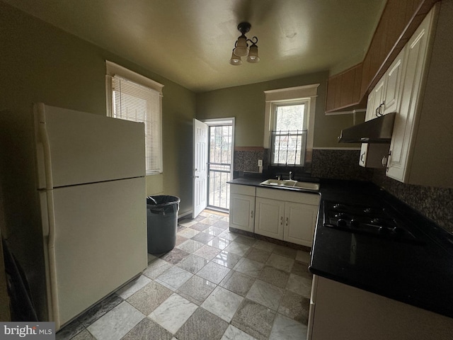 kitchen featuring decorative backsplash, dark countertops, freestanding refrigerator, range hood, and a sink