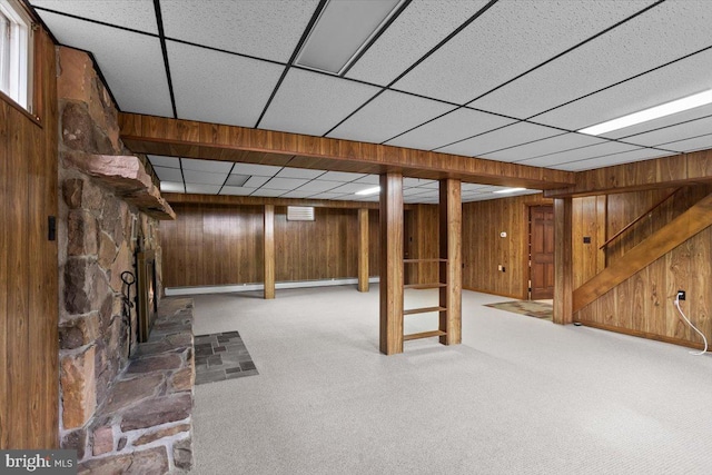 basement with carpet floors, a stone fireplace, a paneled ceiling, and wooden walls