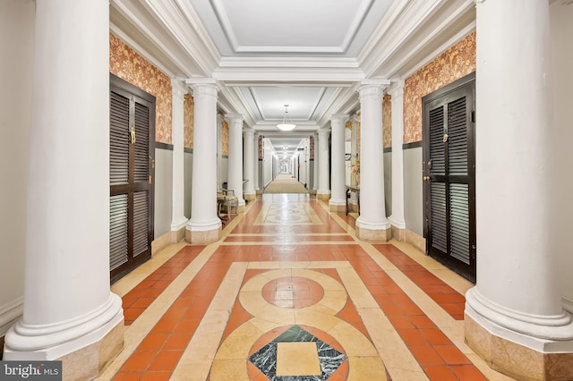 hall with a tray ceiling, crown molding, ornate columns, tile patterned flooring, and baseboards