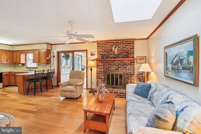 living area with a skylight, light wood finished floors, a ceiling fan, ornamental molding, and a brick fireplace