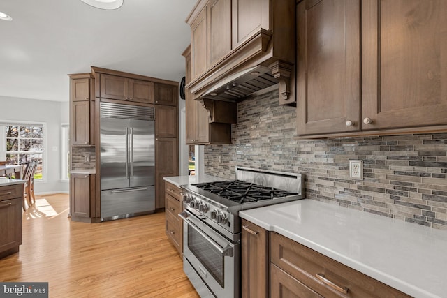 kitchen with premium range hood, light wood-type flooring, high quality appliances, backsplash, and light countertops