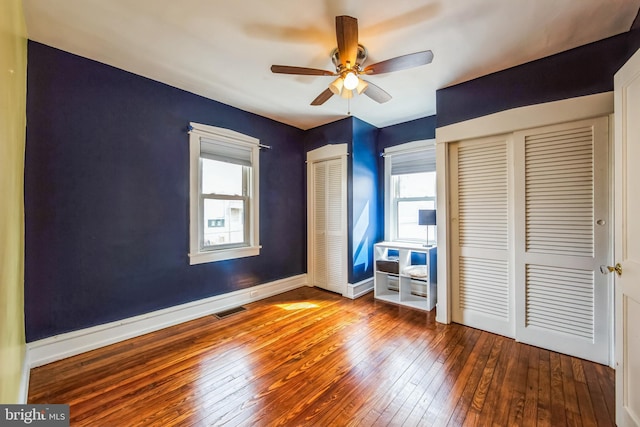 unfurnished bedroom with two closets, visible vents, a ceiling fan, baseboards, and hardwood / wood-style flooring