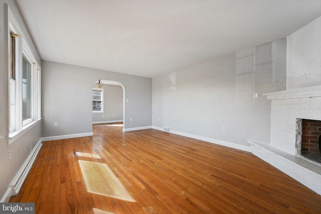 unfurnished living room with a baseboard heating unit, arched walkways, plenty of natural light, and a stone fireplace