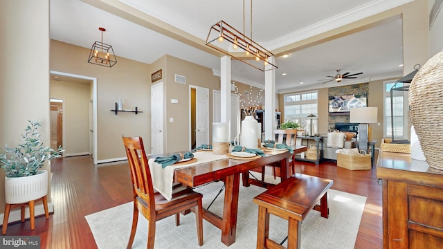 dining area featuring wood finished floors, a stone fireplace, ornamental molding, and ceiling fan with notable chandelier