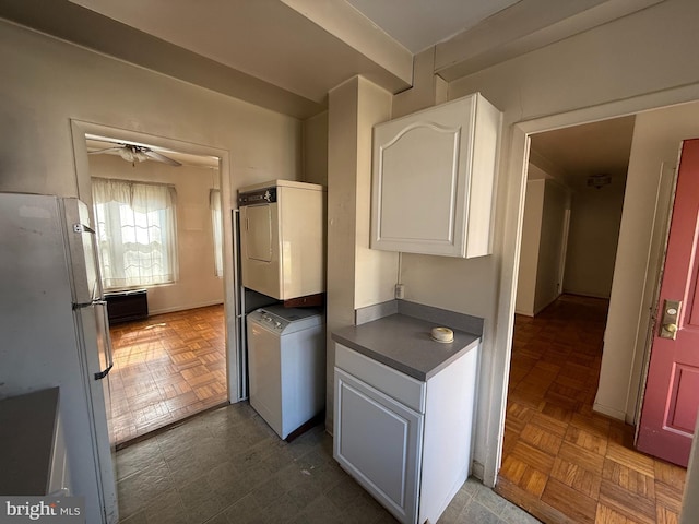 kitchen featuring dark countertops, ceiling fan, freestanding refrigerator, stacked washing maching and dryer, and white cabinetry