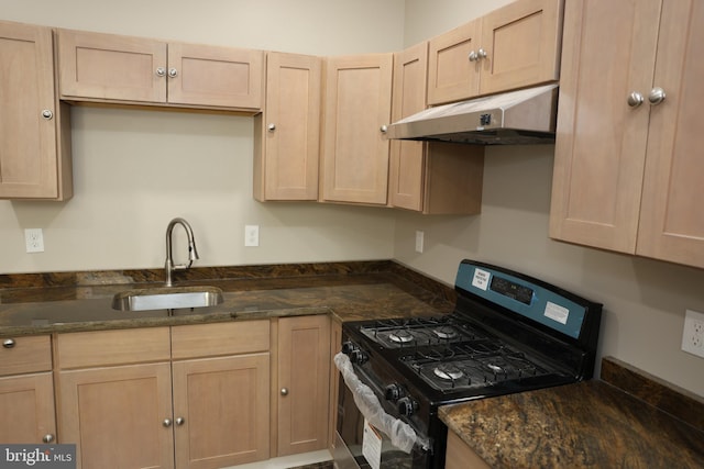 kitchen with under cabinet range hood, light brown cabinetry, gas range, dark stone counters, and a sink