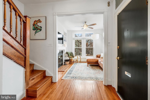 foyer with light wood-style flooring, a ceiling fan, baseboards, stairs, and a glass covered fireplace