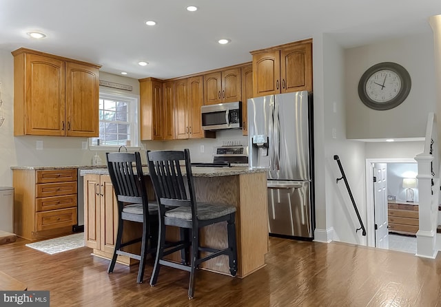 kitchen featuring brown cabinetry, dark wood-style flooring, a center island, light stone countertops, and stainless steel appliances