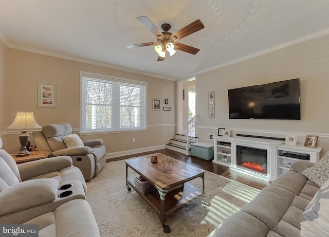 living area with baseboards, a ceiling fan, a glass covered fireplace, ornamental molding, and wood finished floors