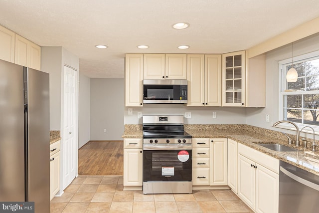 kitchen featuring cream cabinets, stainless steel appliances, a sink, light stone countertops, and glass insert cabinets