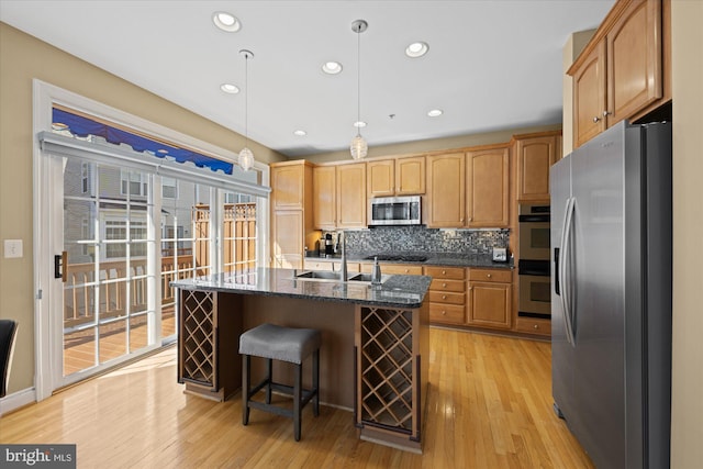 kitchen with light wood finished floors, a kitchen island with sink, a sink, appliances with stainless steel finishes, and backsplash