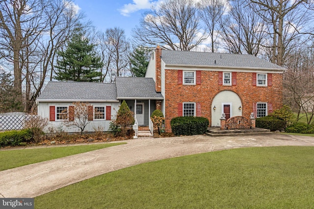 colonial home with driveway, a shingled roof, a chimney, a front lawn, and brick siding