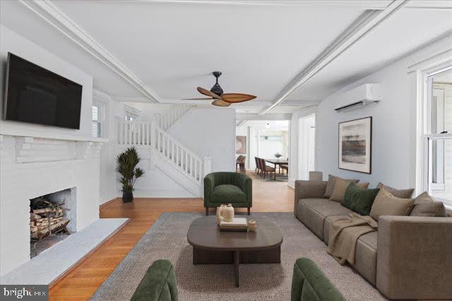 living room with a wall unit AC, a fireplace with raised hearth, stairway, ceiling fan, and wood finished floors